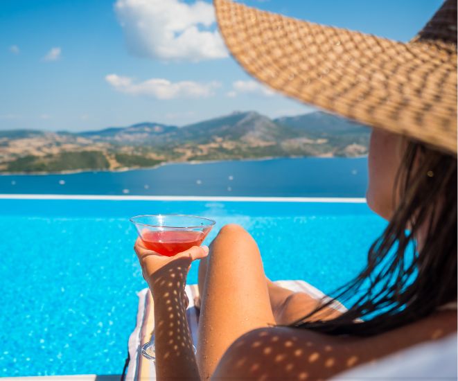 A woman wearing a straw hat and holding a red drink. She is sitting in front of clear blue water.