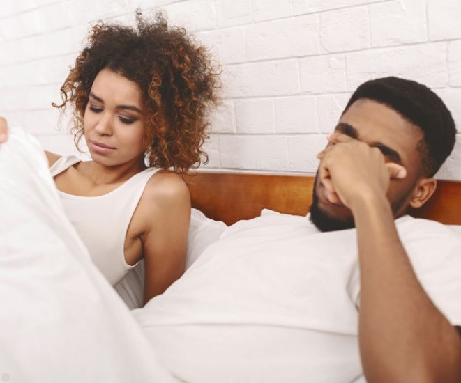 A couple in bed together. The woman is looking under the blanket, unimpressed. The man is hiding his face in his hands, embarrassed.