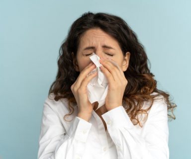 A woman blowing her nose, in front of a blue background.